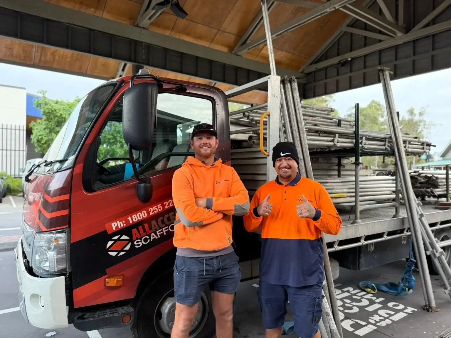 Two workers in high-vis orange shirts standing in front of an All Access Hire truck loaded with scaffolding equipment, under a covered work area.