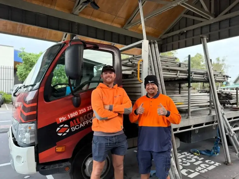 Two workers in high-vis orange shirts standing in front of an All Access Hire truck loaded with scaffolding equipment, under a covered work area.