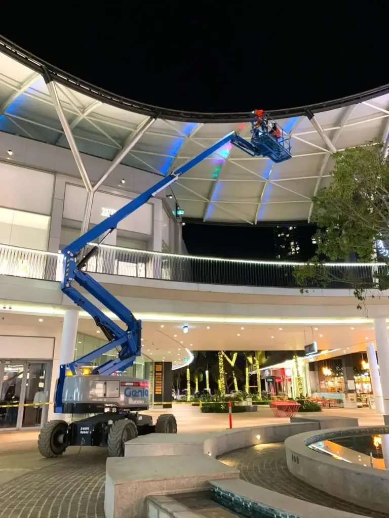 Genie Z-60 FE articulated boom lift operating at night under canopy at Pacific Fair, used for elevated maintenance or installation tasks.